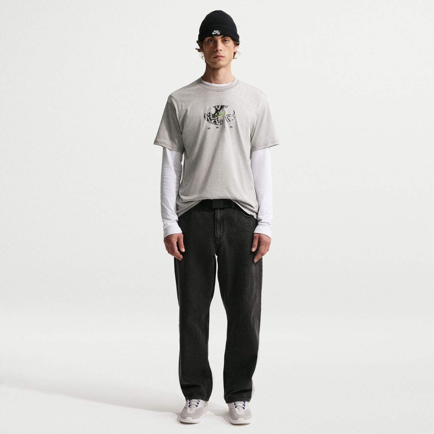 Person wearing a light gray t-shirt with a logo, black pants, and a black cap on a white background