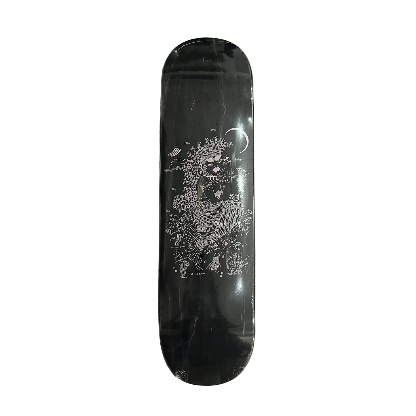 A black skateboard deck with a detailed white and gray illustration of a mermaid under a crescent moon, resting on a white surface.