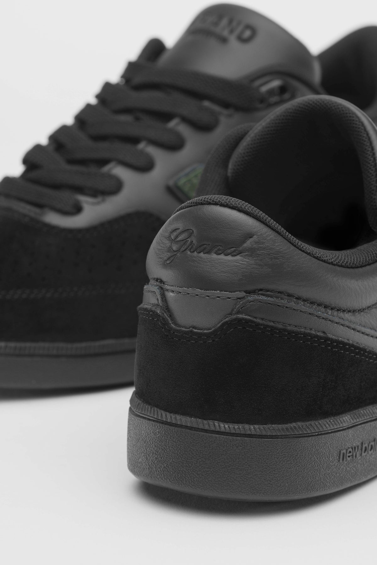 A close-up studio shot of black New Balance sneakers, showing the heel and side profile of the foreground shoe with the brand name 'Grand' embossed on the heel counter.