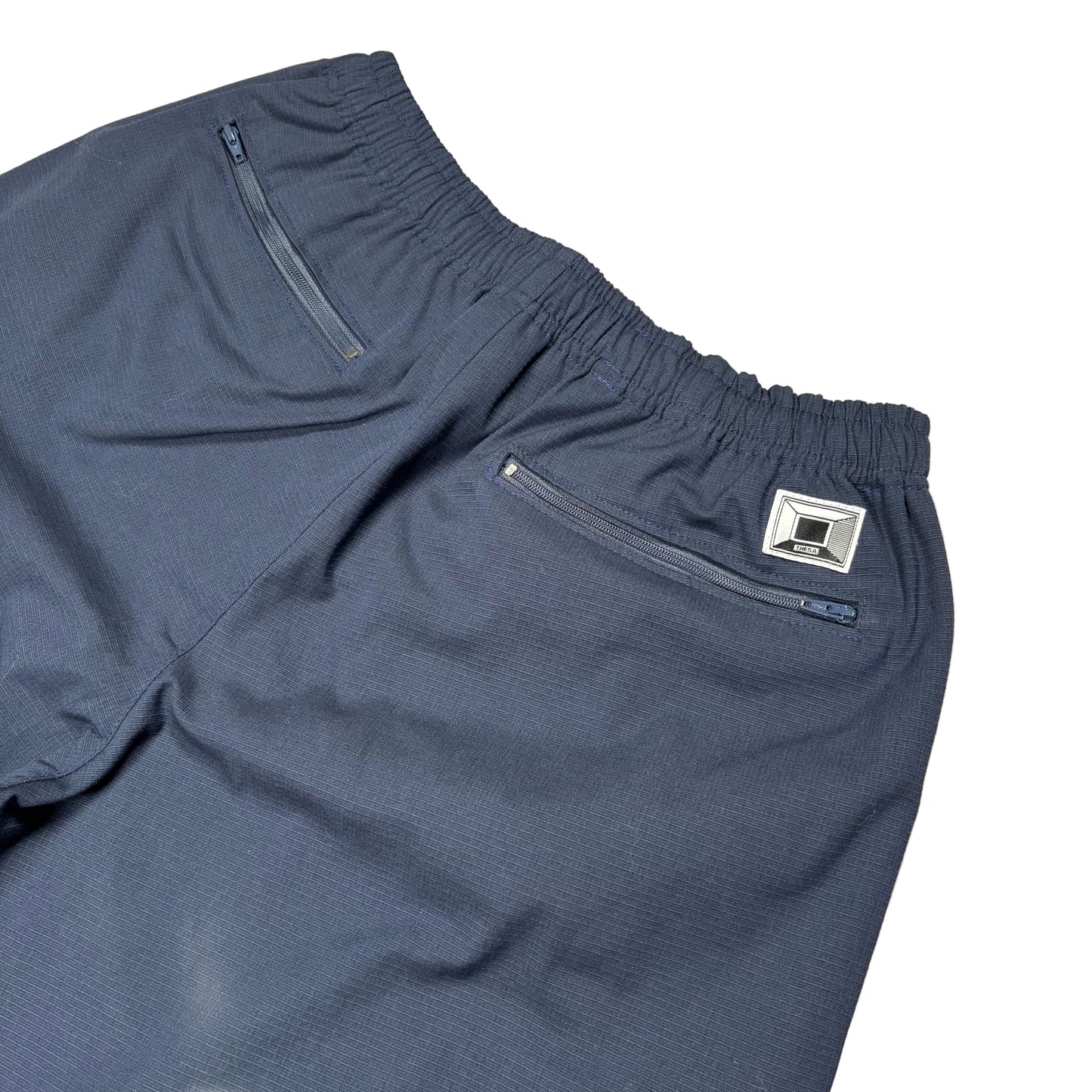 Close-up of dark slate blue athletic shorts showing the elastic waistband, a zippered side pocket, and a small silver brand patch on a white background.