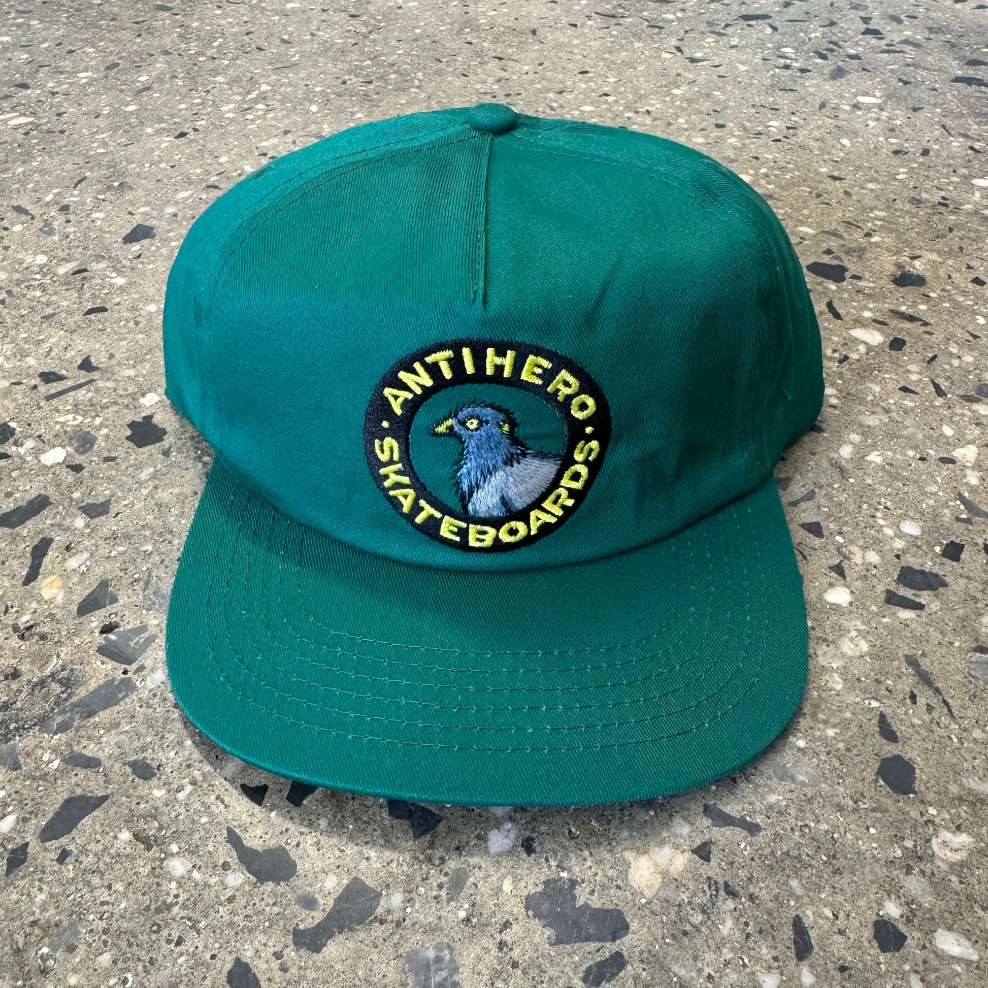 Green hat with yellow text and black circle embroidery with pigeon in the center of it