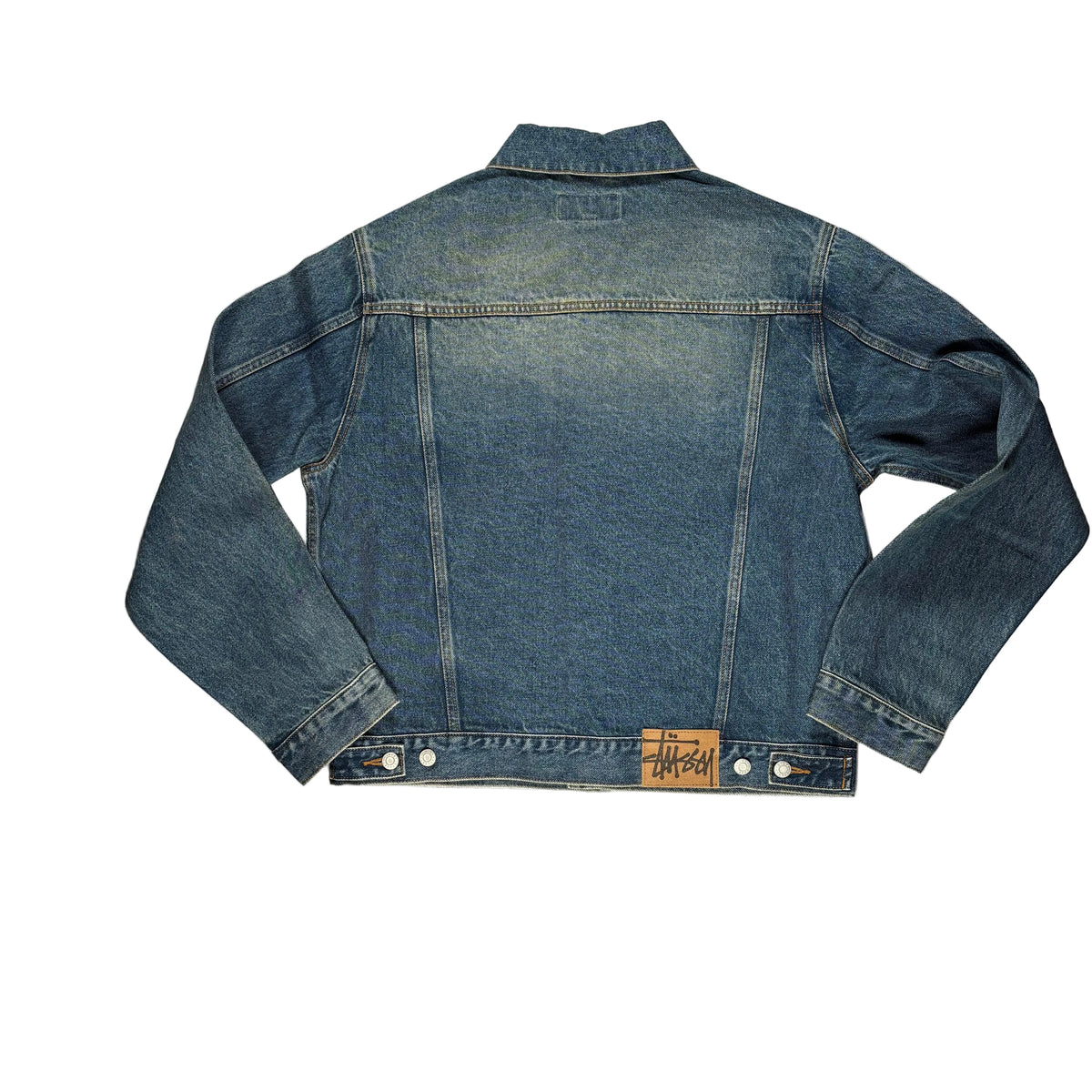 A blue denim jacket laid flat on a white surface, photographed from above. The jacket is shown from the back with long sleeves, contrast stitching, and a small tan leather patch near the hem.
