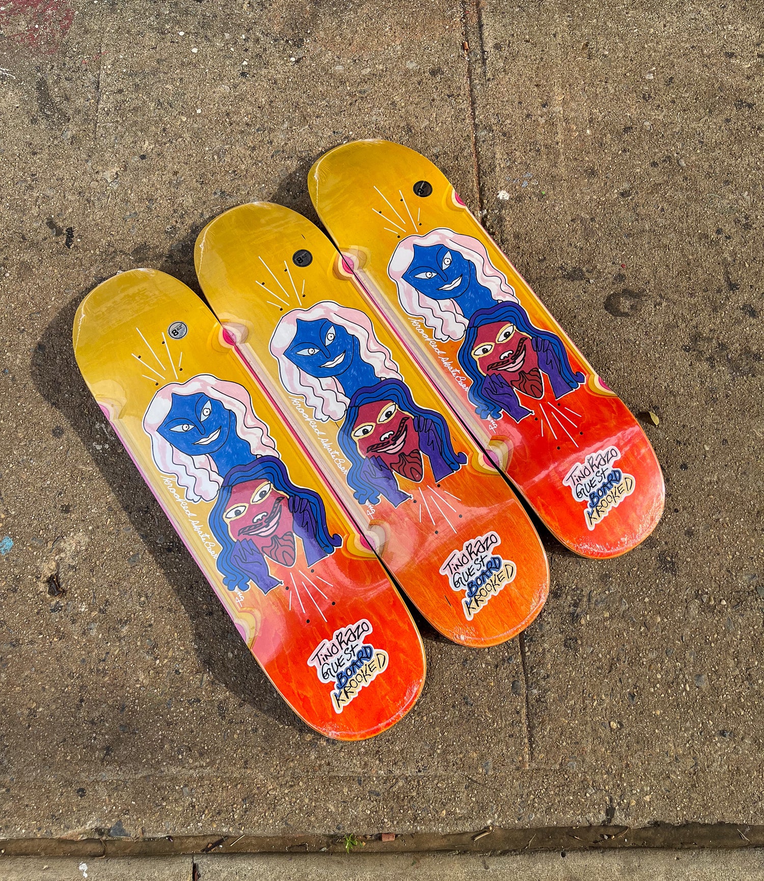Krooked skateboard decks with yellow and red wood grain faded stain and a multi colored graphic in the middle with text on the tail of the board