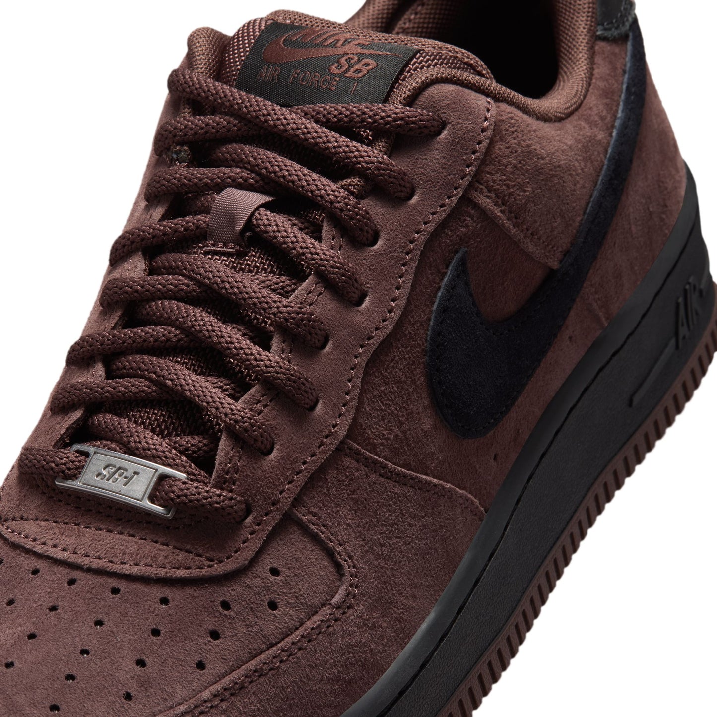 A close-up of a Nike Air Force 1 Low sneaker in dark brown suede and black, featuring matching brown laces, a black Swoosh, and a metallic silver 'SB-1' plate on the eyelet.