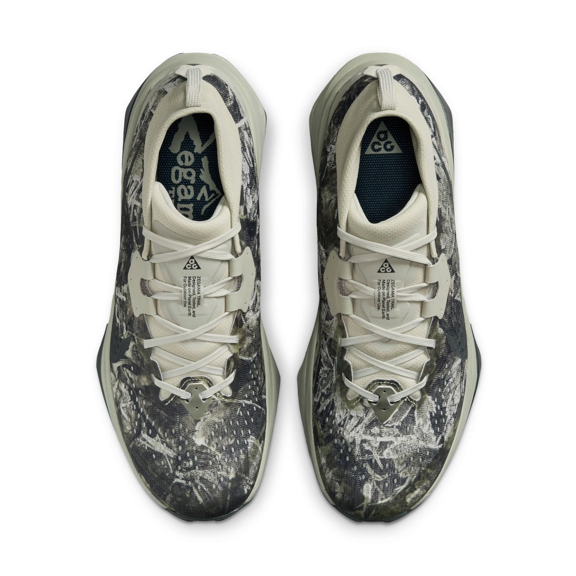 A pair of Nike zegama trail sneakers shown from a top-down view. The shoes feature a dark gray and black abstract camo pattern on a light gray and off-white base, with white laces