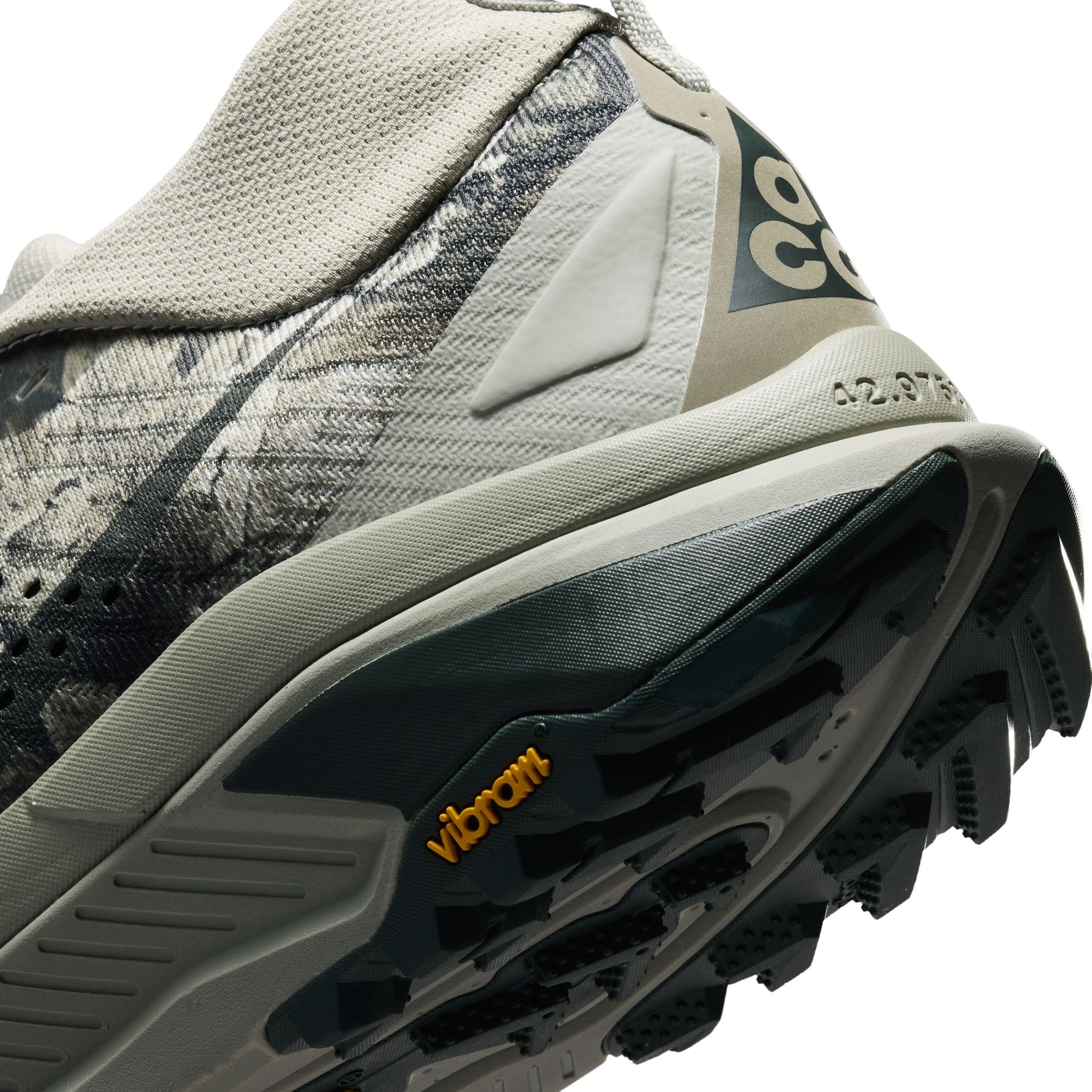 Close-up of a trail running shoe showing the midsole, outsole, heel counter, and part of the upper in gray, black, and beige tones with a visible Vibram logo.