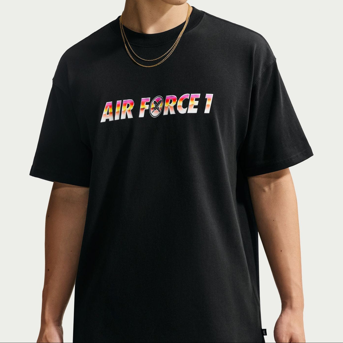 A model wearing a black t-shirt with 'AIR FORCE 1' printed across the chest in a colorful gradient pattern, styled with gold necklaces and red reflective sunglasses against a light gray background.