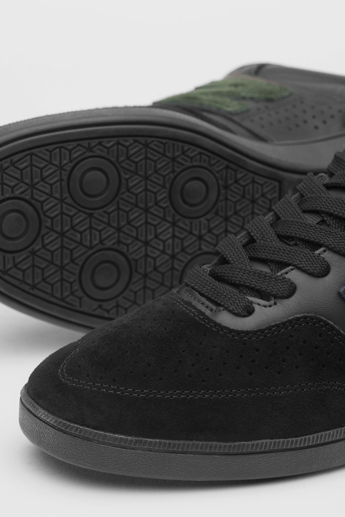 Close-up of a black suede sneaker with black laces and a black sole, with another shoe shown from the back displaying its textured black outsole.