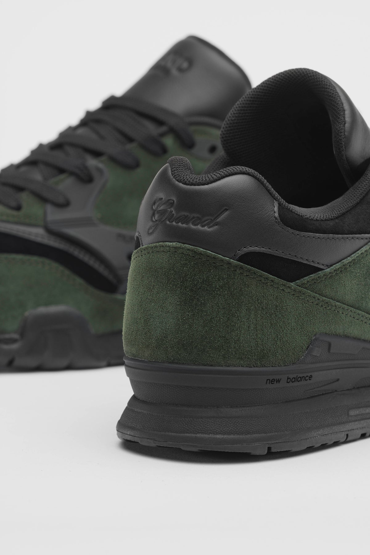 Close-up of a New Balance sneaker in dark green suede and black leather, showing the heel, midsole, and embossed 'Grand' branding.