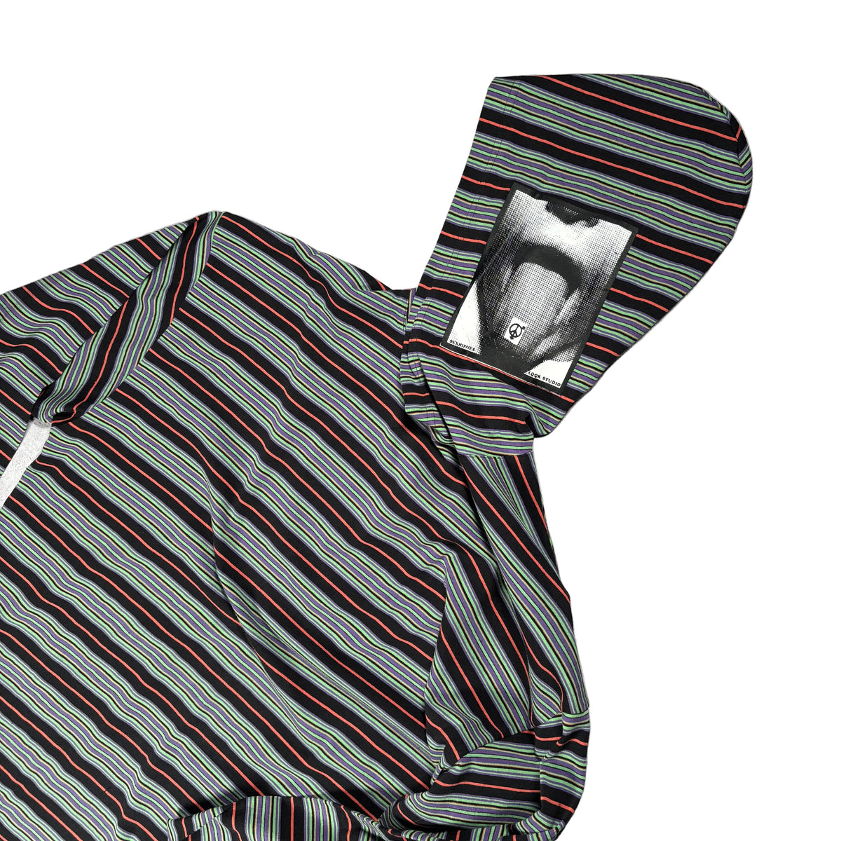 A hooded striped long-sleeve shirt laid flat on a white surface. The fabric has diagonal stripes in gray, black, red, and thin blue lines. The hood has a patch of a mouth with the tongue sticking out.