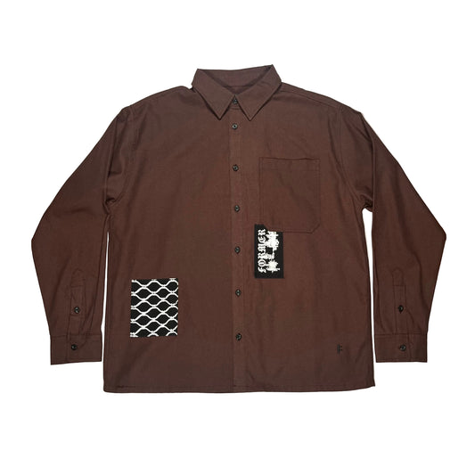 A dark brown long-sleeve button-up shirt laid flat on a white surface. The shirt features a chest pocket, black buttons, a black-and-white geometric patterned patch on the lower left front, and a black brand patch on the right front pocket area.