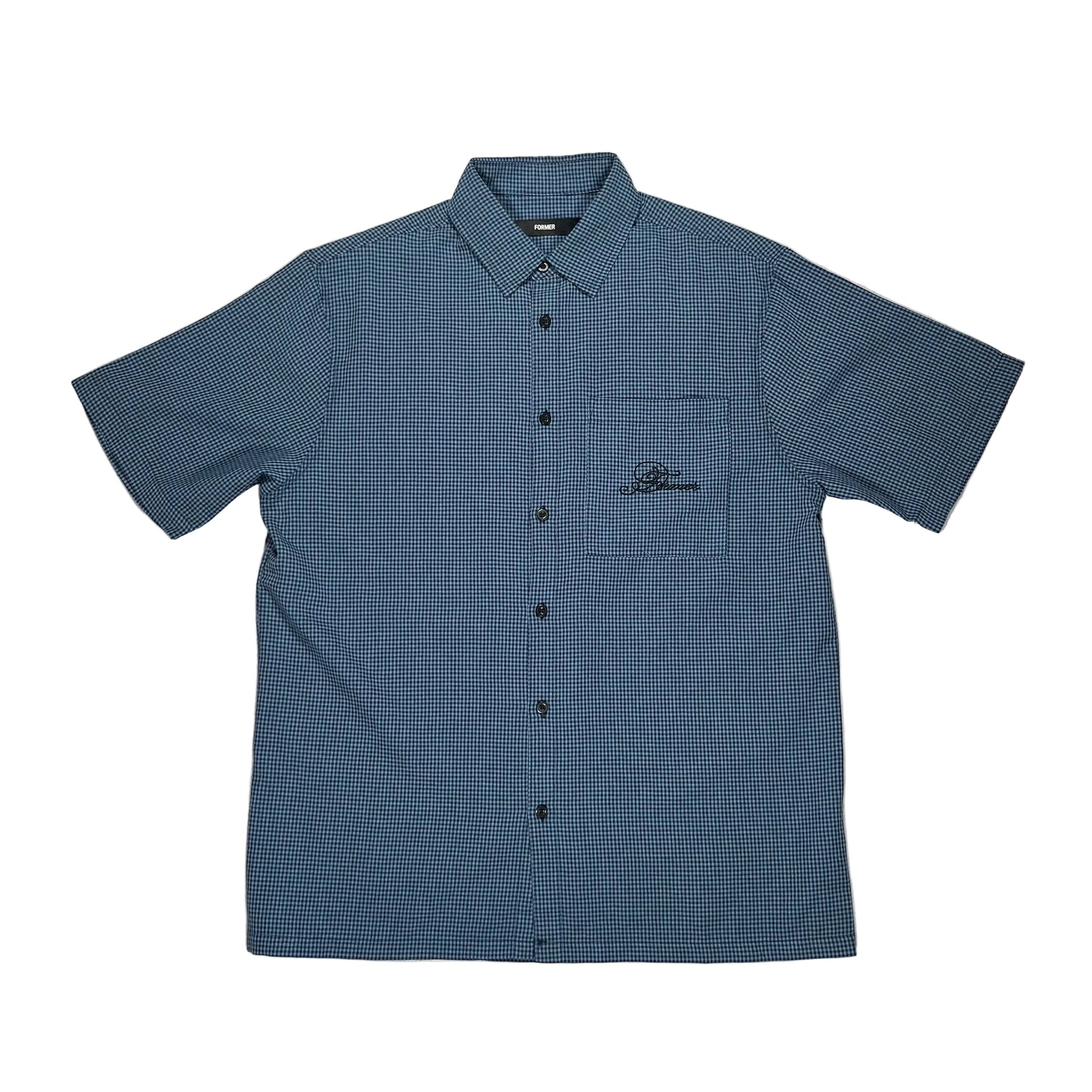 A short-sleeve button-up shirt in a blue and black micro-check pattern, laid flat on a white suface. The shirt features a chest pocket with a black embroidered logo on the left chest, and is photographed from a top-down.