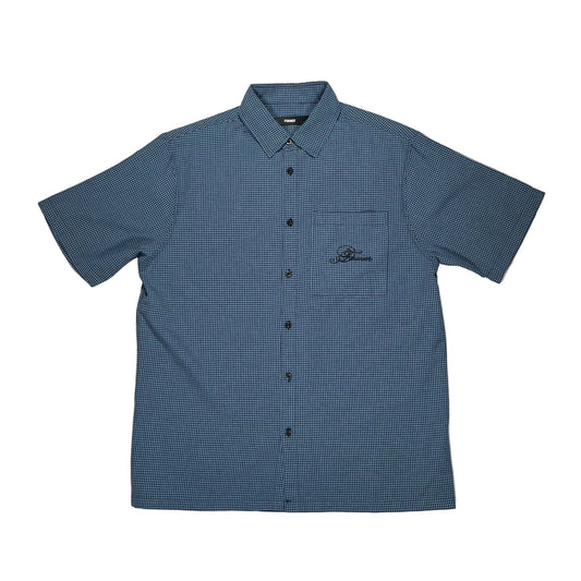 A short-sleeve button-up shirt in a blue and black micro-check pattern, laid flat on a white suface. The shirt features a chest pocket with a black embroidered logo on the left chest, and is photographed from a top-down.