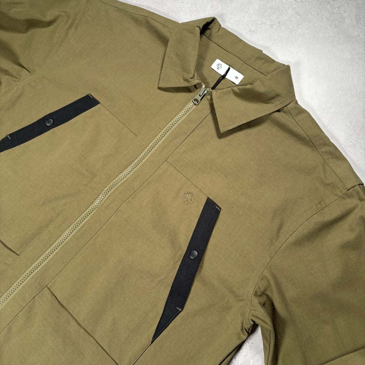 An olive green utility jacket with black contrast panels and a front zipper, laid flat on a white surface.