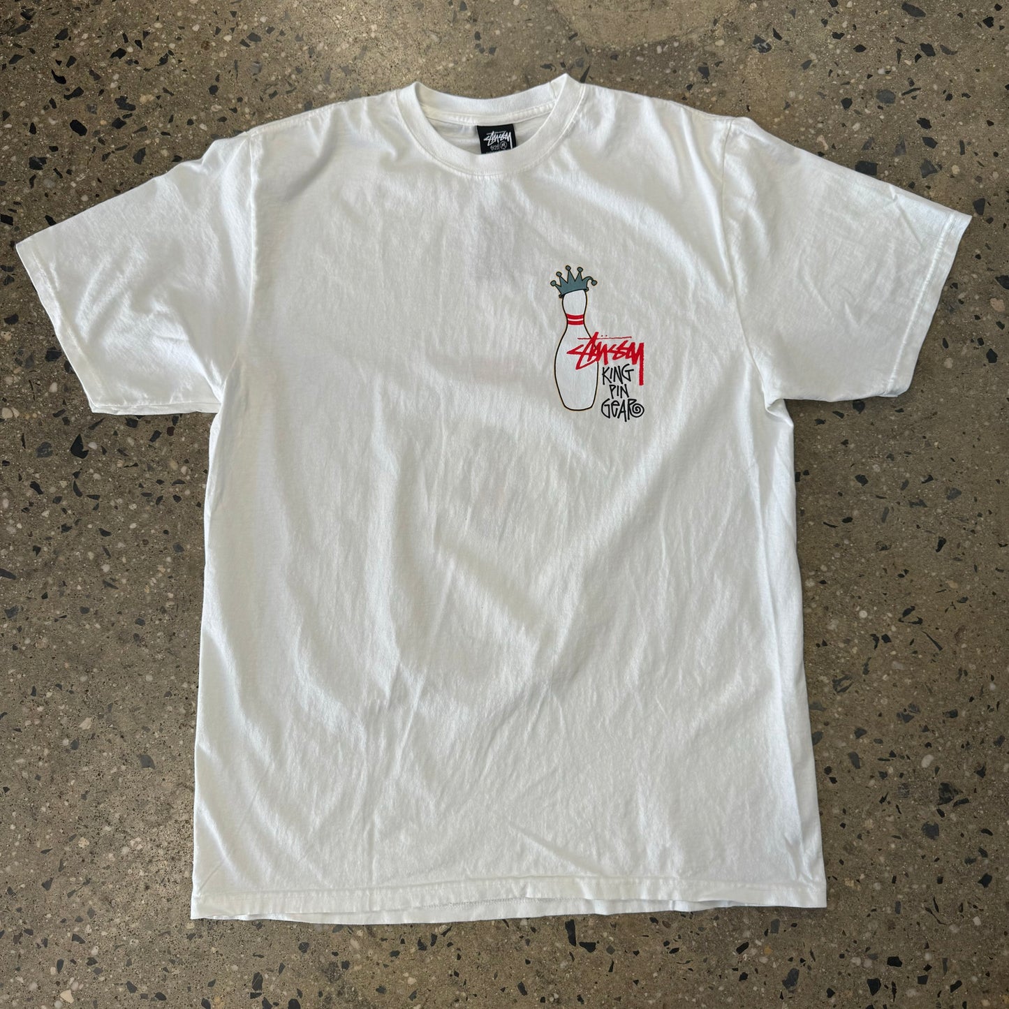 front view of natural white stussy teeshirt with small bowling pin on left chest wearing a crown