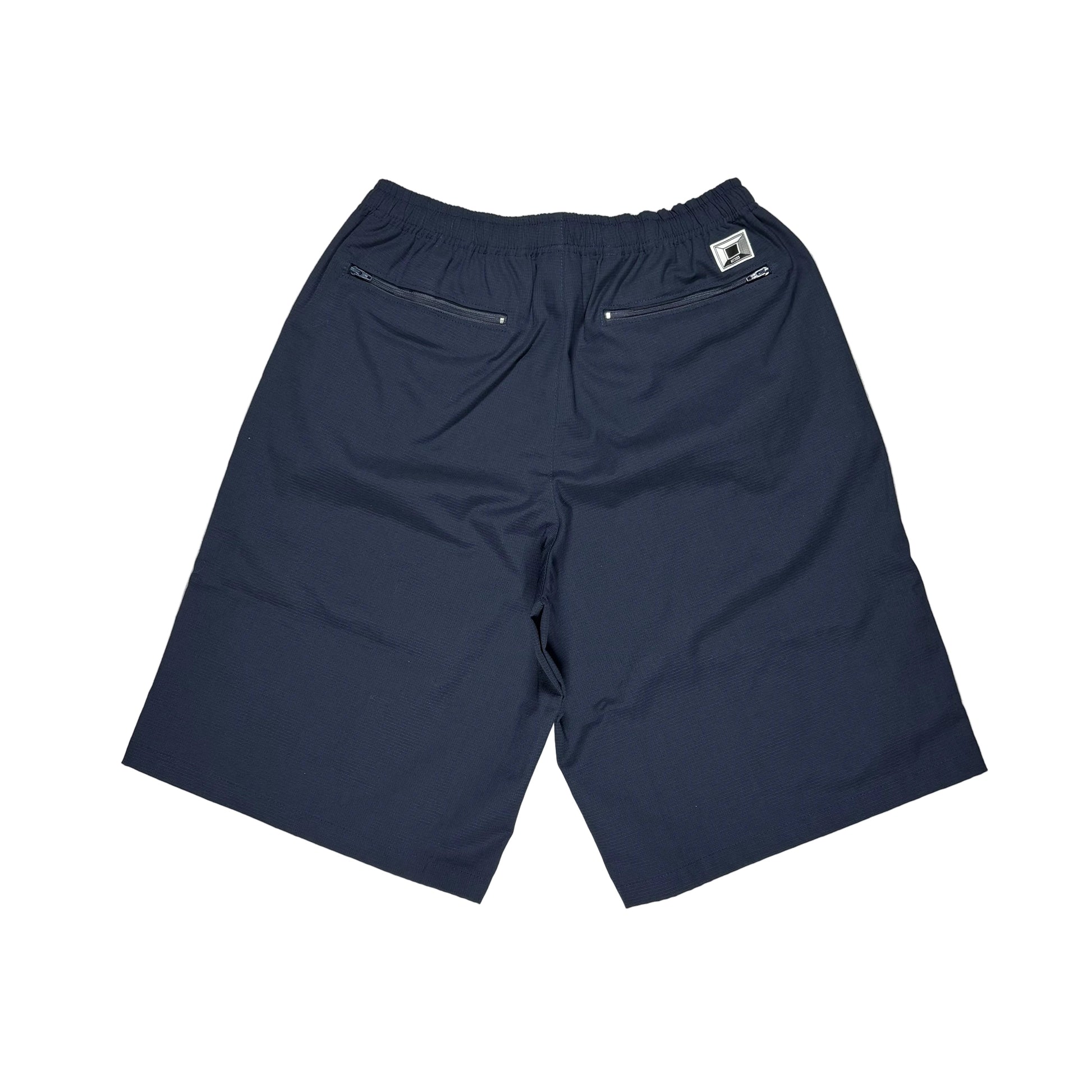 A pair of navy blue athletic shorts laid flat on a white surface, showing the back waistband, zippered pockets, and a small white brand tag. Photographed from behind