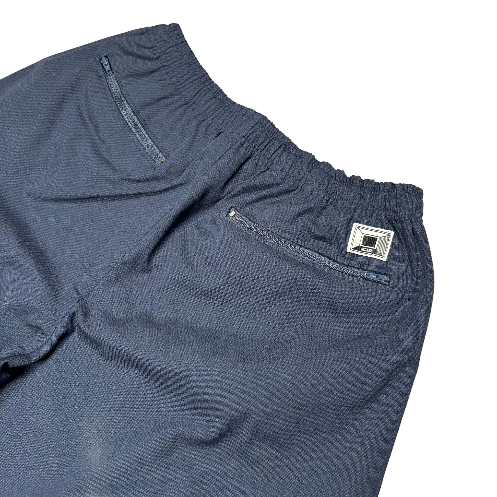 Close-up of dark slate blue athletic shorts showing the elastic waistband, a zippered side pocket, and a small silver brand patch on a white background.