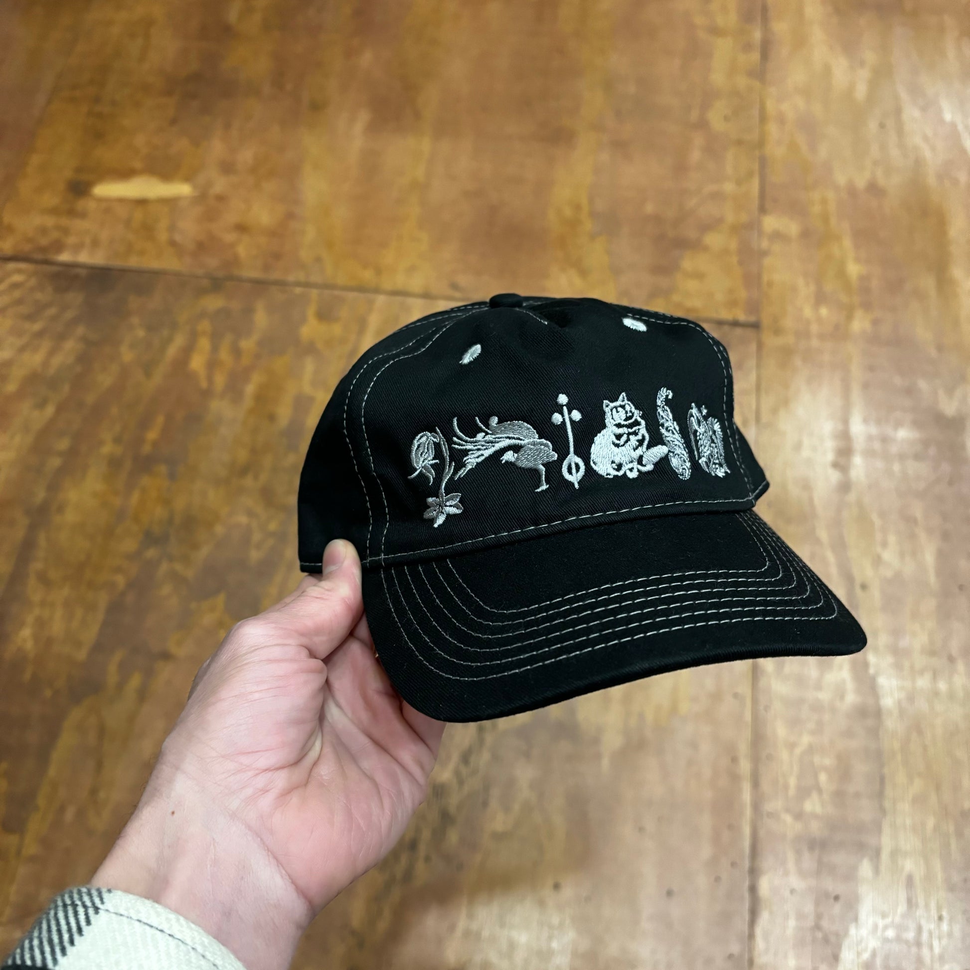 front view of black hat with white stitching, and embroidered plants and animals on front