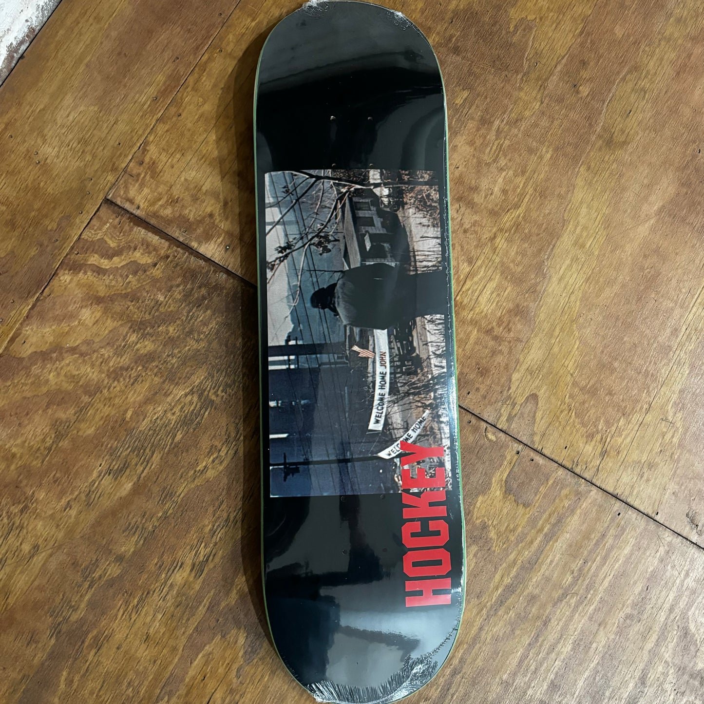 horizontal graphic on bottom of skateboard deck with photo of banner saying welcome home john on it