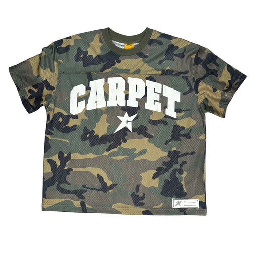 A short-sleeve crewneck jersey in a green, brown, and black camouflage pattern, featuring the word 'CARPET' in bold white arched letters with a white star underneath, laid flat on a white surface