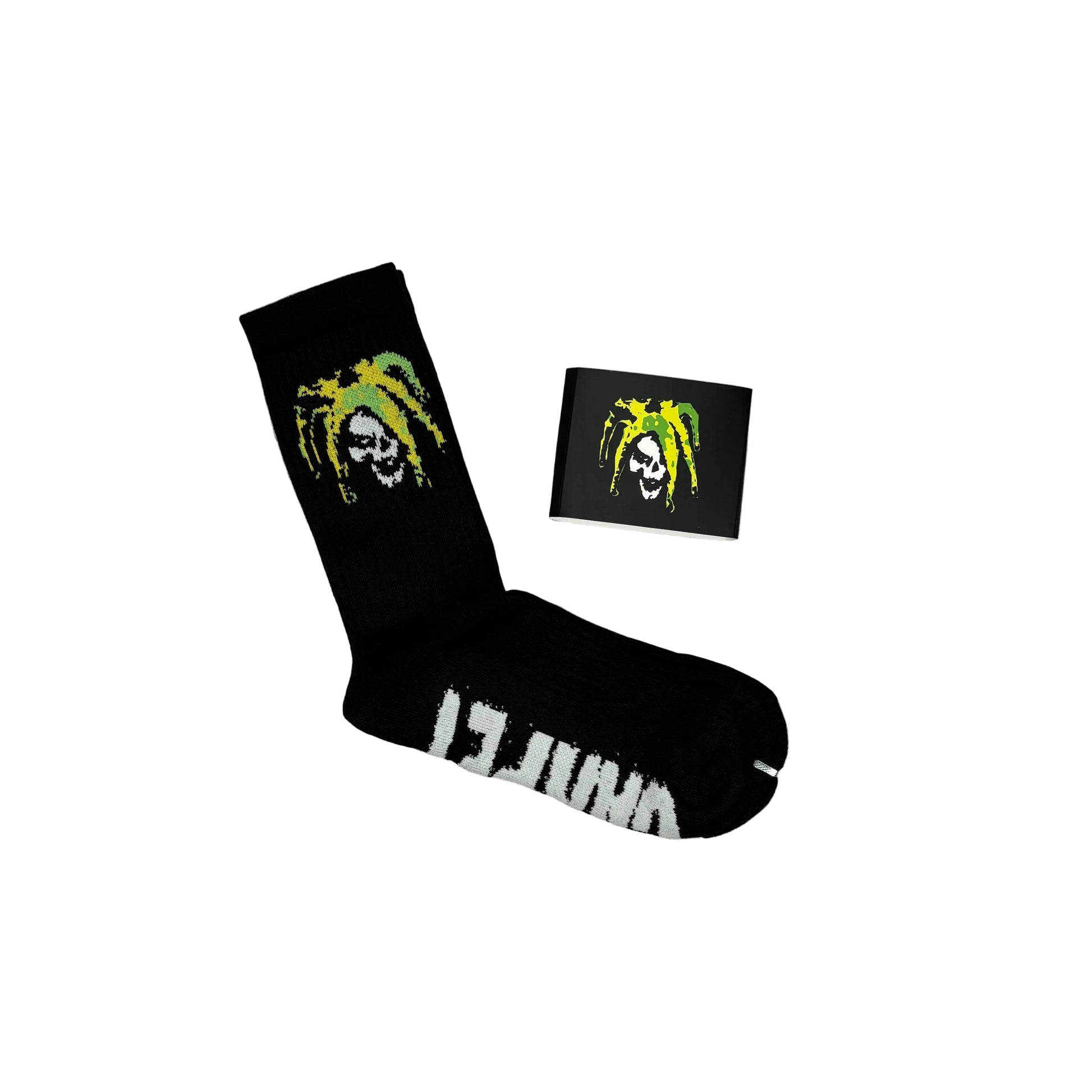 A pair of black crew socks with a green and white skull-and-dreadlock graphic, shown on a white background with one sock laid flat and a paper sock packaging sleeve with the dreadlock skull