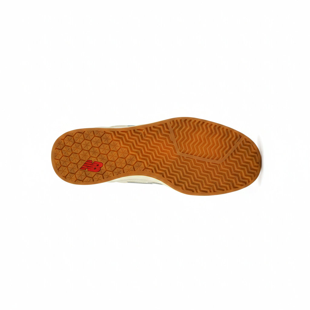 A top-down view of a sneaker's orange gum outsole, featuring a red New Balance logo and a honeycomb pattern, set against white background.