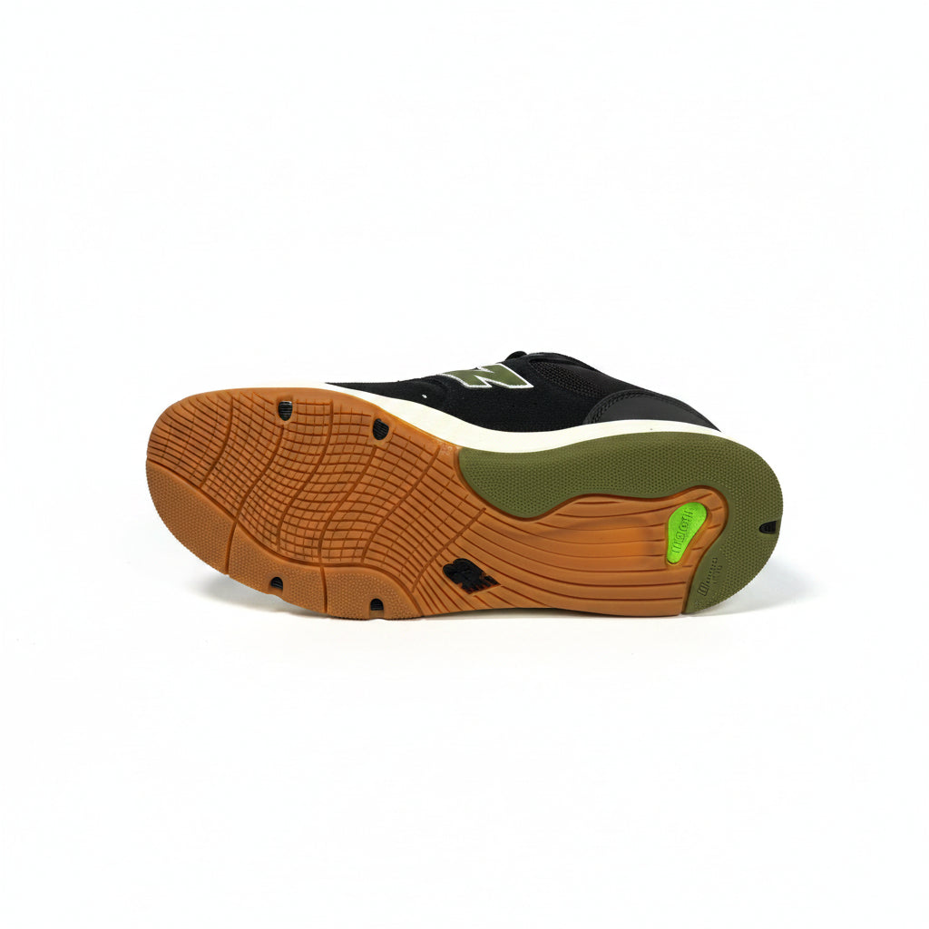 A single New Balance sneaker shown from the bottom, displaying its black suede upper, olive green heel, and a brown rubber outsole with a small bright green accent, on a white background