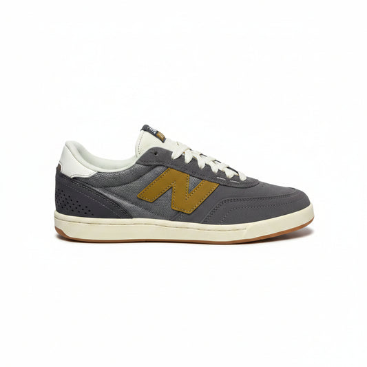 A single New Balance sneaker in dark gray suede with a gold 'N' logo, white laces, and a cream-colored midsole, photographed against a white background