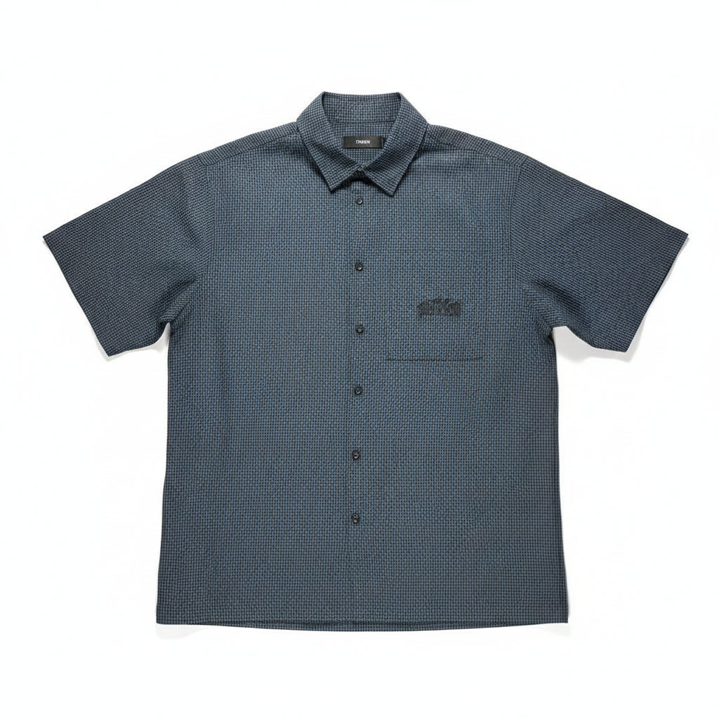 A short-sleeve button-up shirt laid flat on a textured light gray surface, bordered by wooden boards on the left and right. The shirt is dark charcoal gray with a subtle micro-check pattern and features a chest pocket with a black embroidered logo.