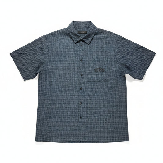 A short-sleeve button-up shirt laid flat on a textured light gray surface, bordered by wooden boards on the left and right. The shirt is dark charcoal gray with a subtle micro-check pattern and features a chest pocket with a black embroidered logo.