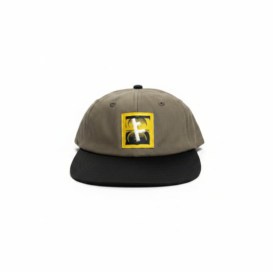 A baseball cap with a brown crown and a black flat brim, featuring a square yellow and black embroidered patch on the front with a white cross symbol.