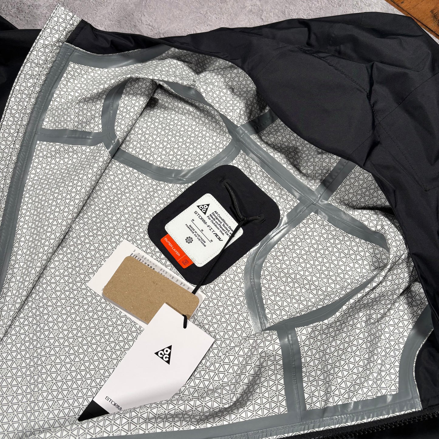 A close-up of a garment laid flat, showing a light gray and white geometric patterned lining with gray trim, a black outer section, and attached tags including a black fabric label with a white triangle logo.