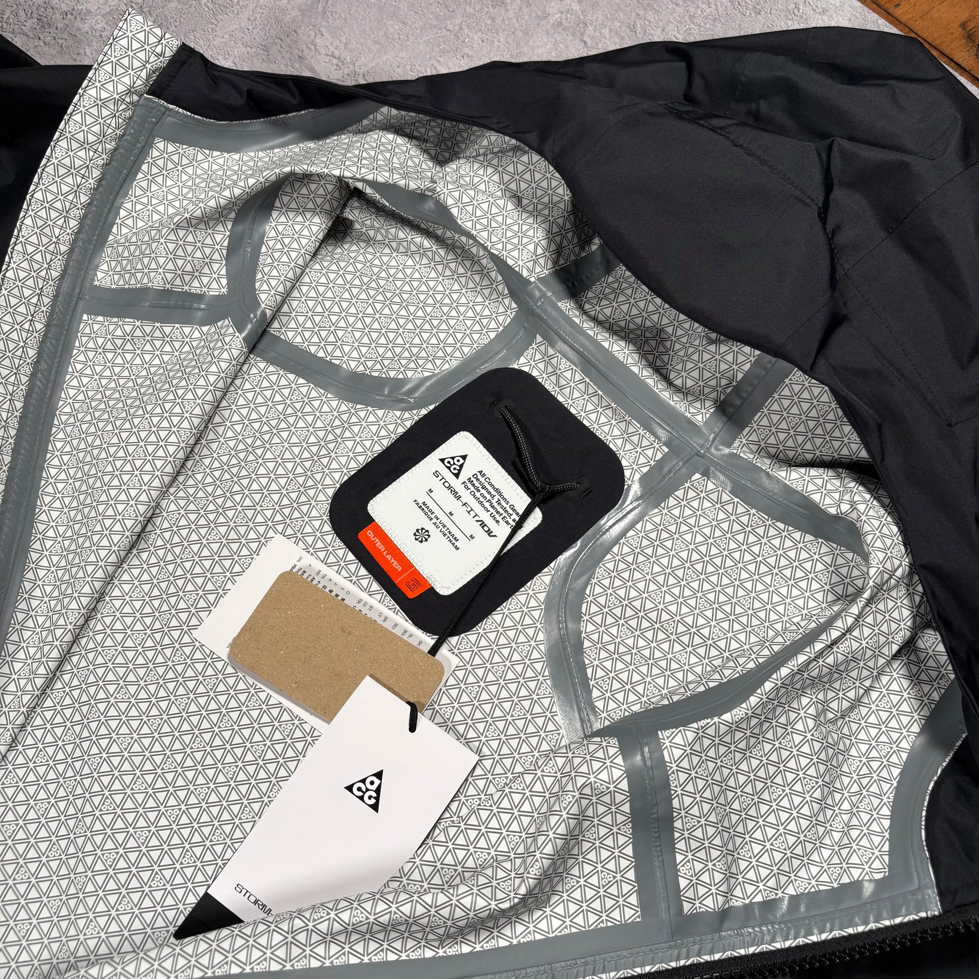A close-up of a garment laid flat, showing a light gray and white geometric patterned lining with gray trim, a black outer section, and attached tags including a black fabric label with a white triangle logo.