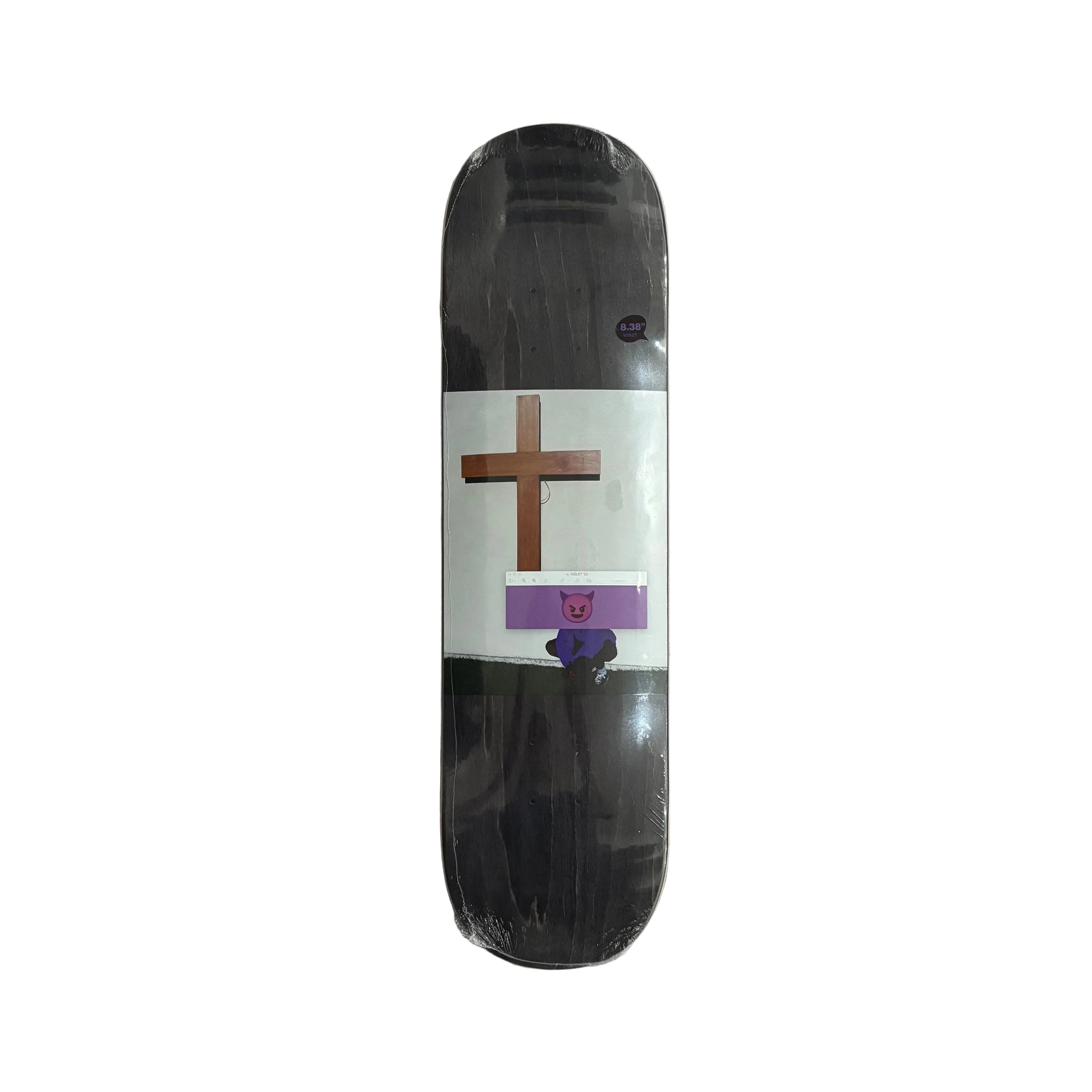 A skateboard deck in its clear plastic shrink wrap, featuring a black and white graphic design with a brown cross and a purple cartoon character, displayed on a white surface.