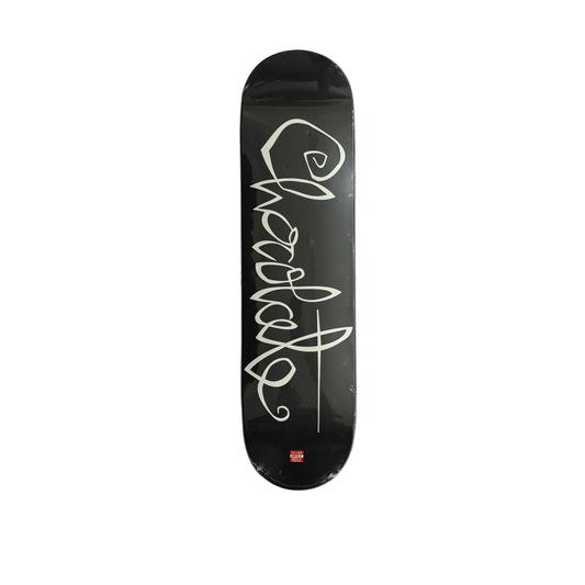 A black skateboard deck with a large white cursive Chocolate graphic and a small red logo near the bottom, photographed from above on a white surface.