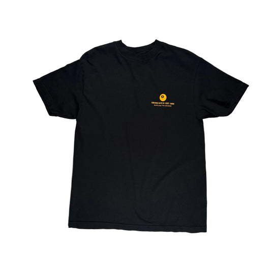 Front side of a black short-sleeve t-shirt laid flat on a white surface, featuring a small gold chest graphic in corner.