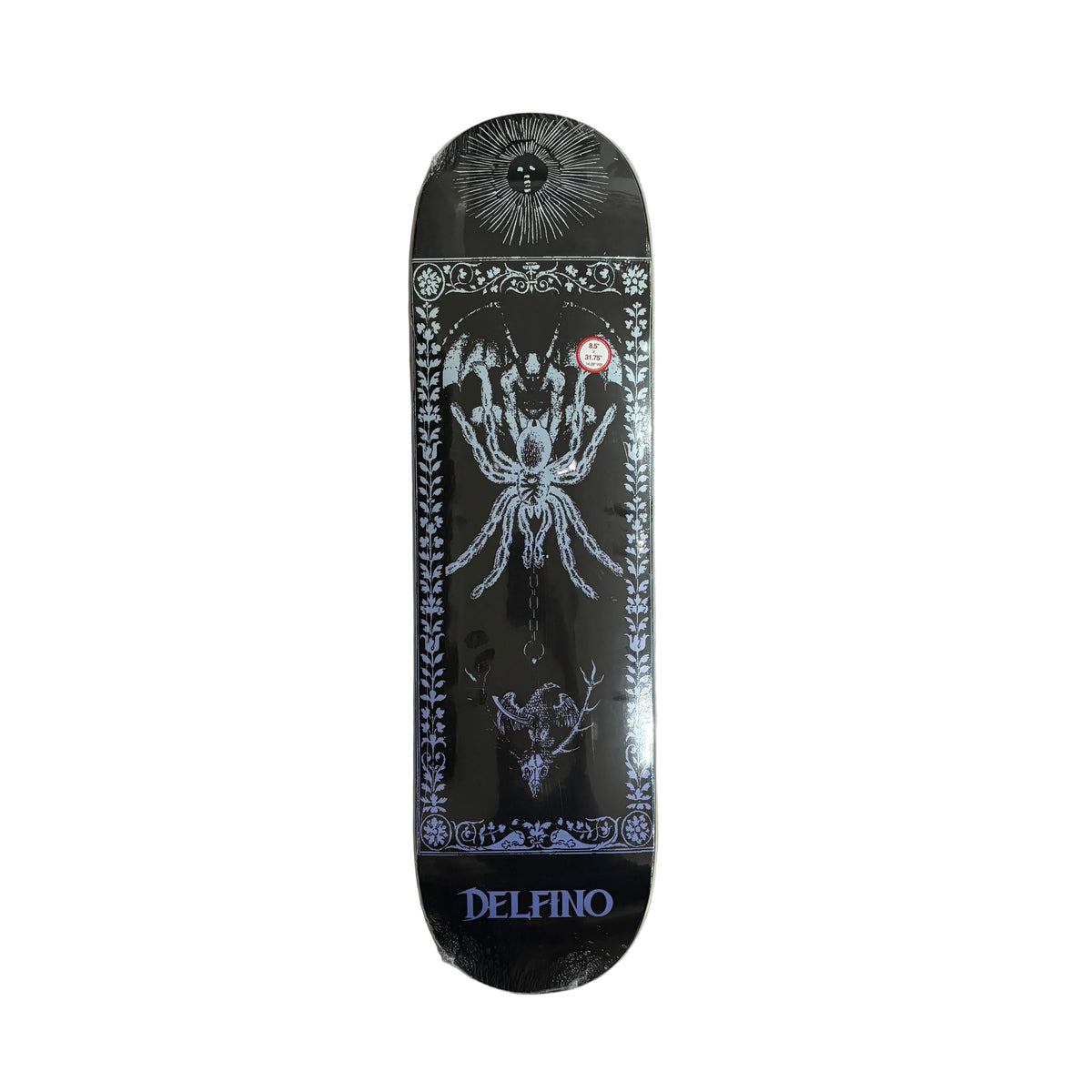 A black Delfino skateboard deck with intricate white and gray gothic illustrations, including a spider, a sun, and a deer skull, resting on a white surface.