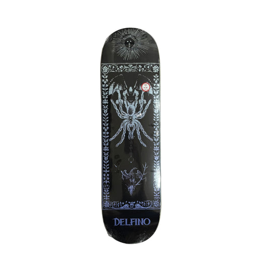 A black Delfino skateboard deck with intricate white and gray gothic illustrations, including a spider, a sun, and a deer skull, resting on a white surface.