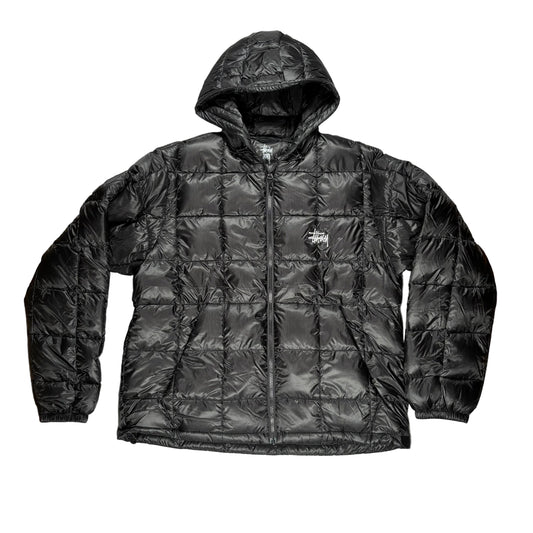 A black quilted puffer jacket with a hood and front zipper, laid flat on a white surface. A small Stussy logo is visible on the left chest.