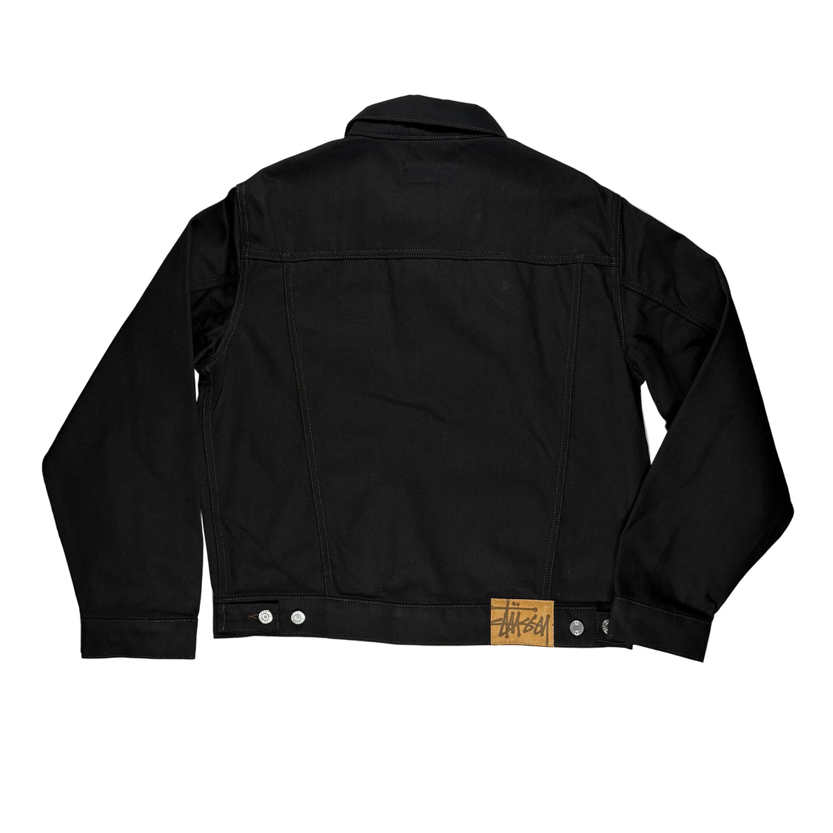 Back side of black denim jacket laid flat on a white surface, with a small tan leather brand patch reading 'Stussy' visible near the waistband.