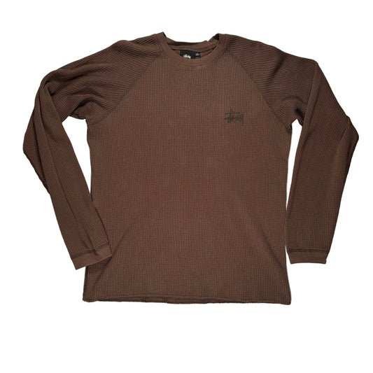 A brown ribbed long-sleeve crewneck shirt by Stüssy, featuring the brand's signature  logo on the left chest, laid flat on a white surface.