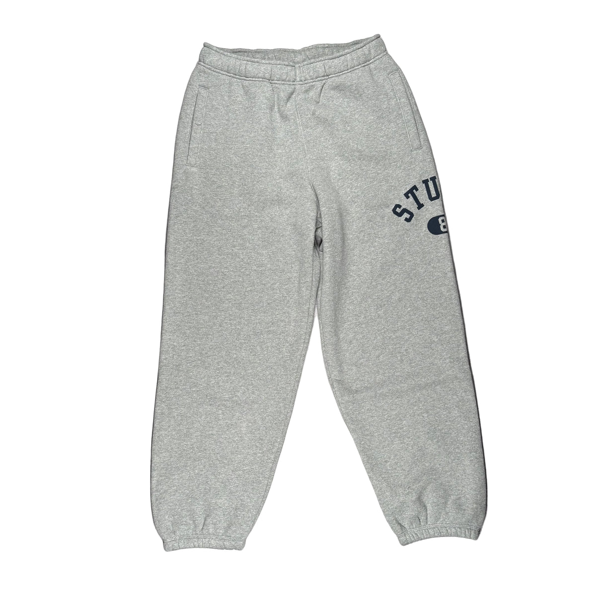 A pair of light heather gray sweatpants laid flat on a white surface. The sweatpants have an elastic waistband, elastic ankle cuffs, and a dark navy blue collegiate-style graphic on the upper left leg that reads 'STUSSY'.