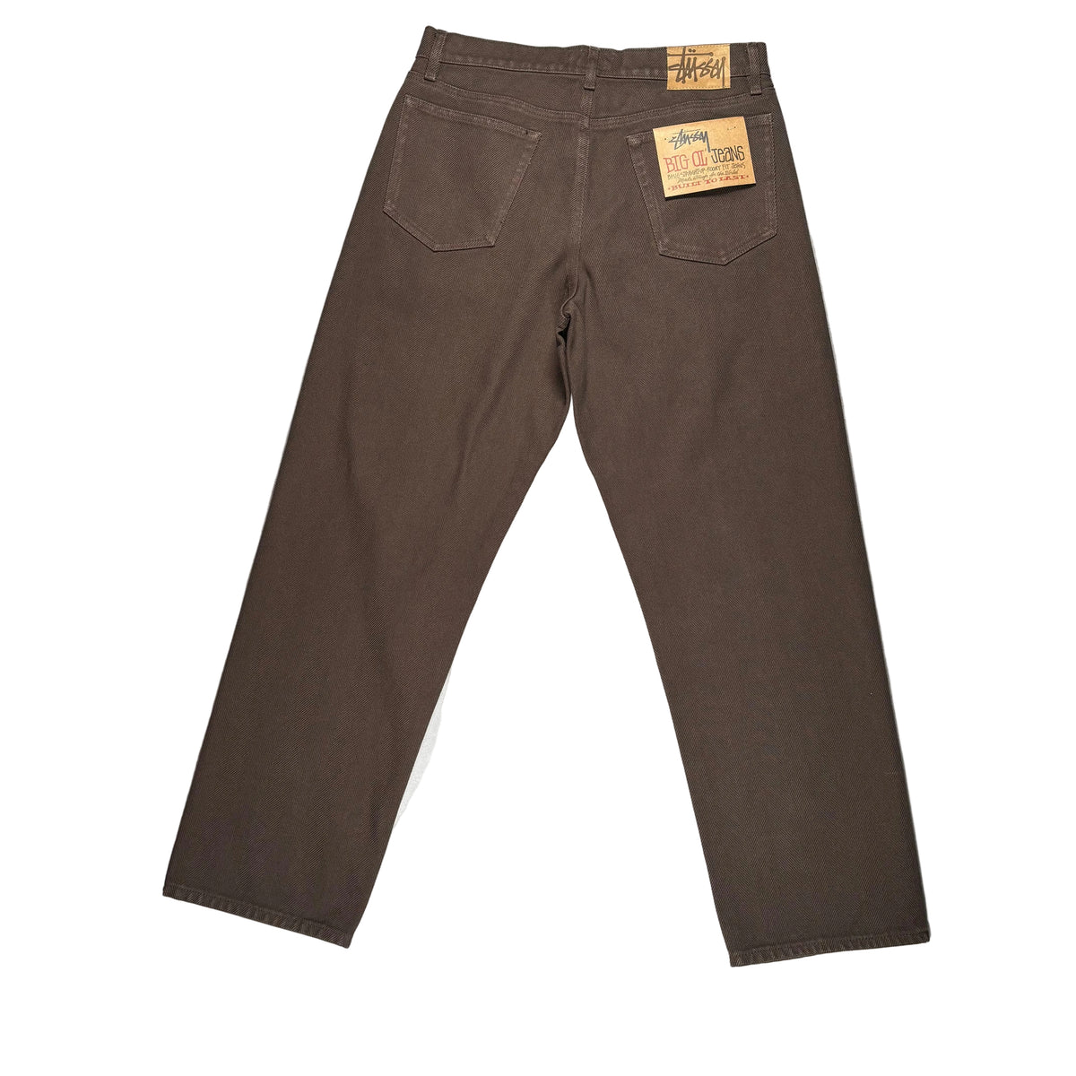 A pair of dark brown jeans laid flat on a white surface. The jeans have a visible leather Stussy patch and a paper tag on the back waistband.