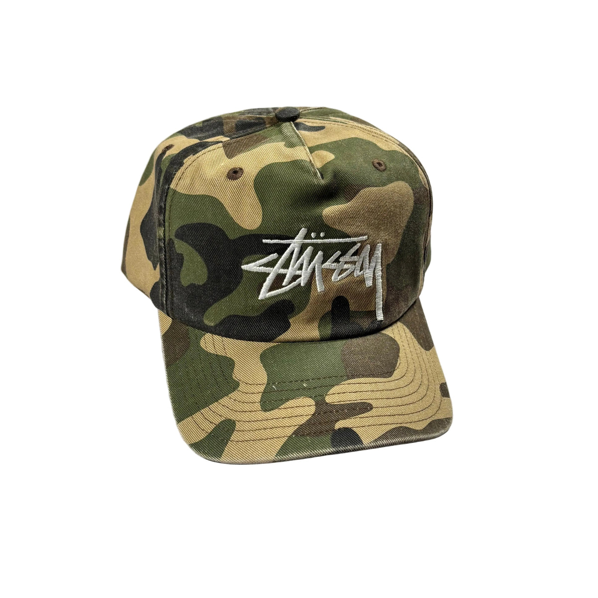 A Stussy baseball cap in a classic camouflage pattern with a white embroidered logo on the front, resting on a white surface.