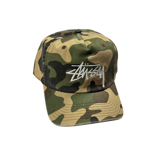 A Stussy baseball cap in a classic camouflage pattern with a white embroidered logo on the front, resting on a white surface.