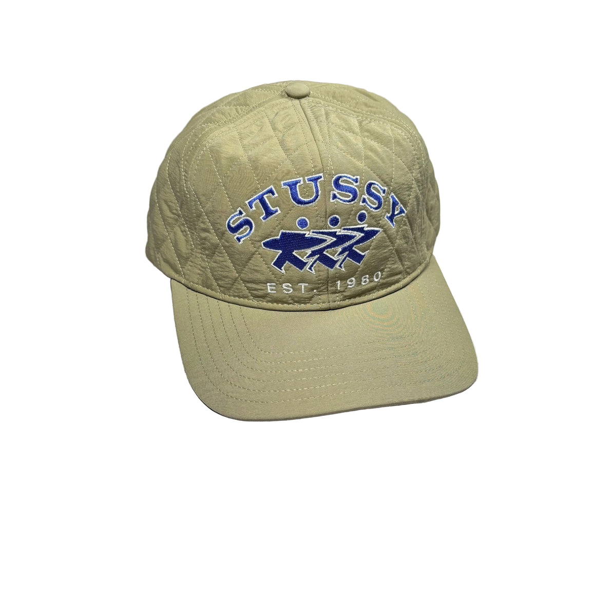 A khaki quilted baseball cap with blue embroidered 'STUSSY' and 'EST. 1980' text on the front, resting on a white surface.