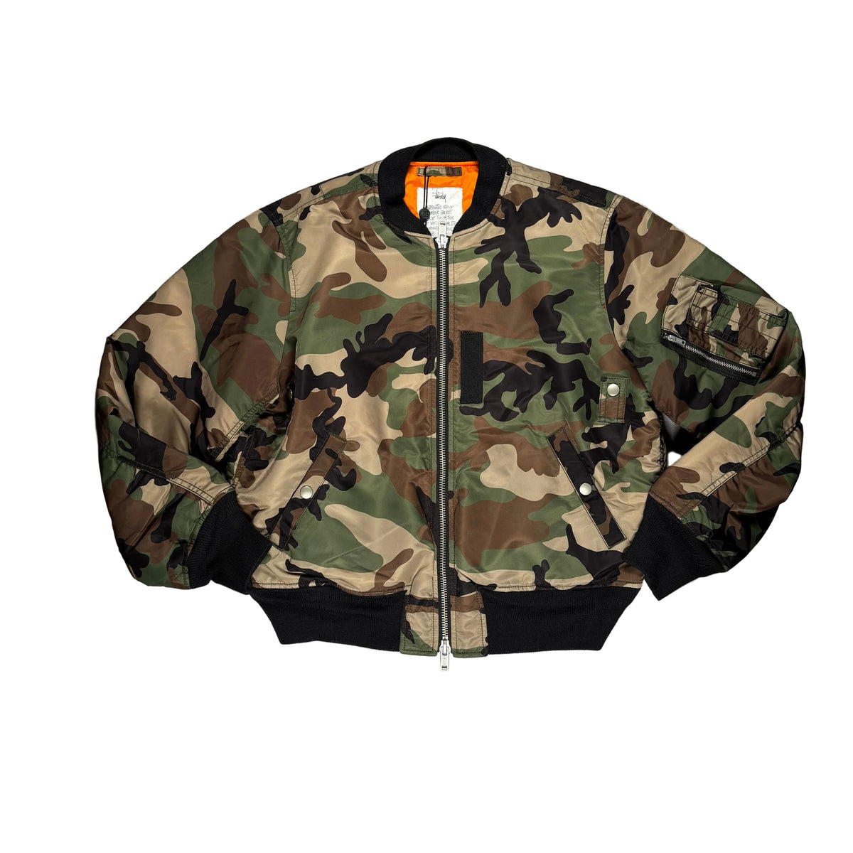 A camouflage bomber jacket laid flat on a white surface. The jacket features a classic woodland camo pattern with green, brown, black, and tan, a black ribbed collar, cuffs, and hem, a front zipper, and a zippered chest pocket.